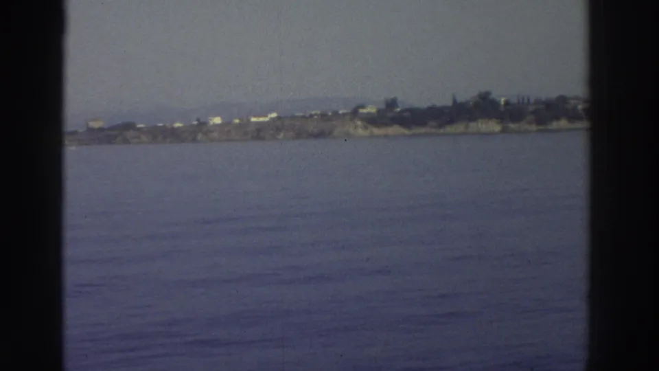 8mm travel footage, c.1976 Greece — wide expanse of Aegean water with a low rocky promontory and scattered white structures on the horizon.