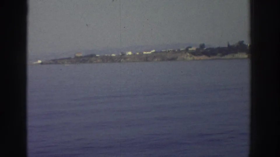 8mm home-movie frame, c.1976 Greece — a flat rocky islet or promontory seen across calm Aegean water from a moving passenger vessel.
