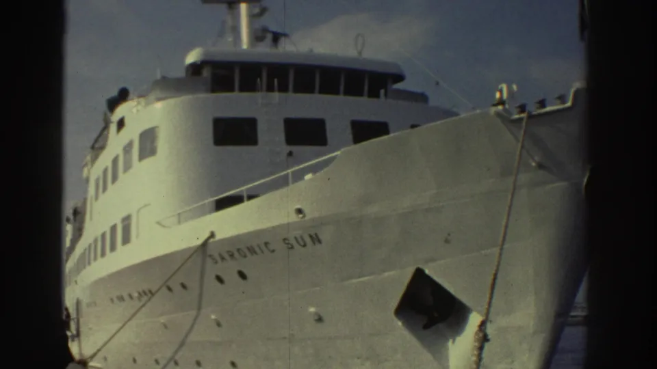 Low-angle 8mm shot of the white cruise ship Saronic Sun moored at a Greek port, circa 1976, hull name clearly legible.