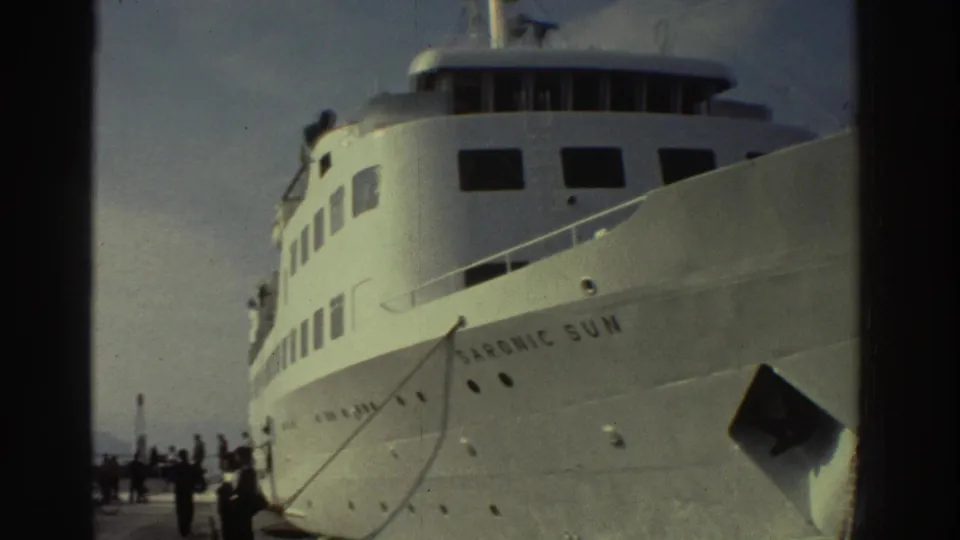 Low-angle 8mm film view of the white Saronic Sun passenger ship at a Greek port with silhouetted passengers on the quay, circa 1976.