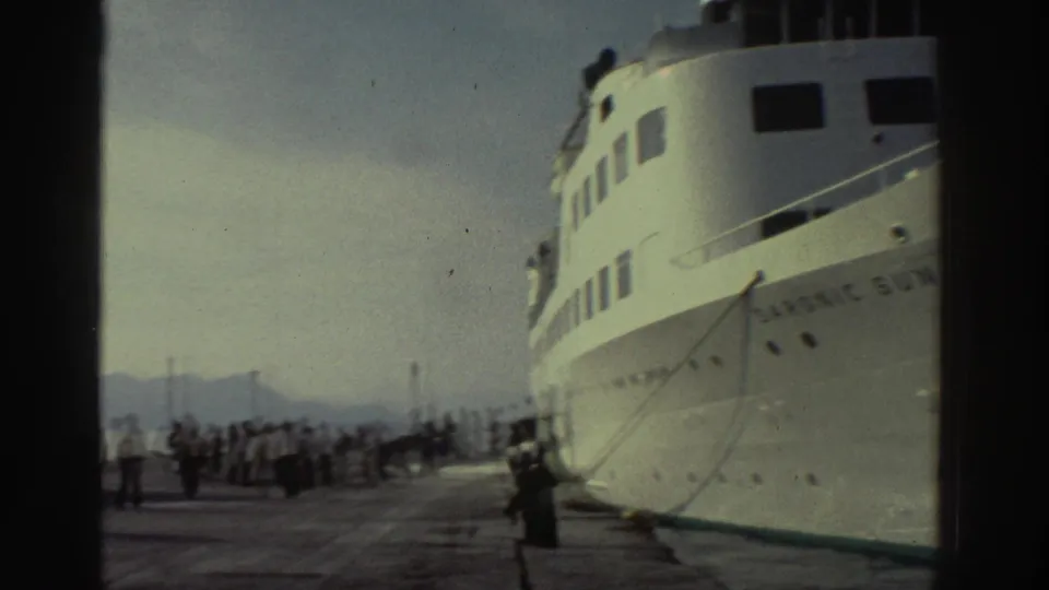 8mm home-movie shot of the Saronic Sun cruise ship bow at a Greek port with quayside figures and distant hills visible, circa 1976.