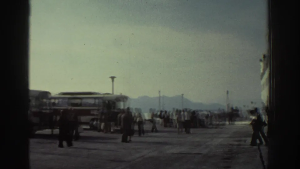 Wide 8mm shot of a Greek port embarkation area with passengers, a terminal building, lamp posts, and hazy distant mountains, circa 1976.
