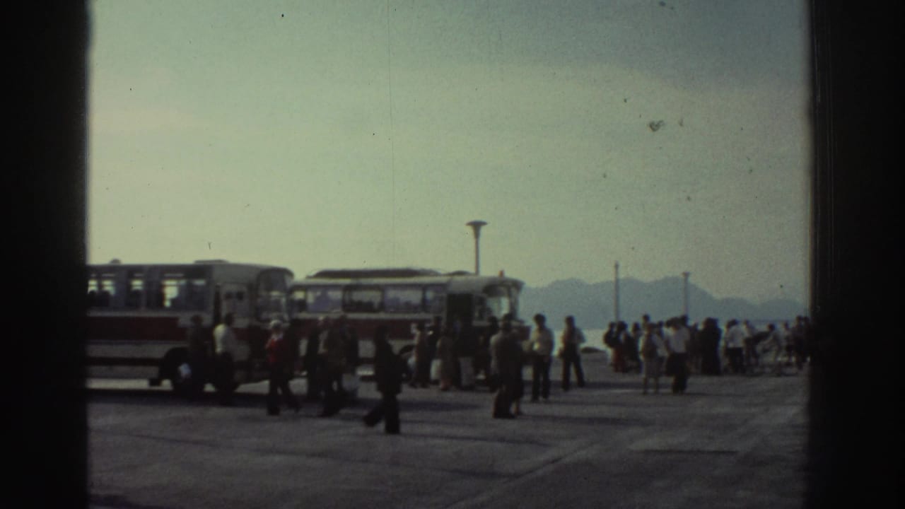 Crowded ferry pier with period buses and passengers milling about, hazy mountains visible in background.