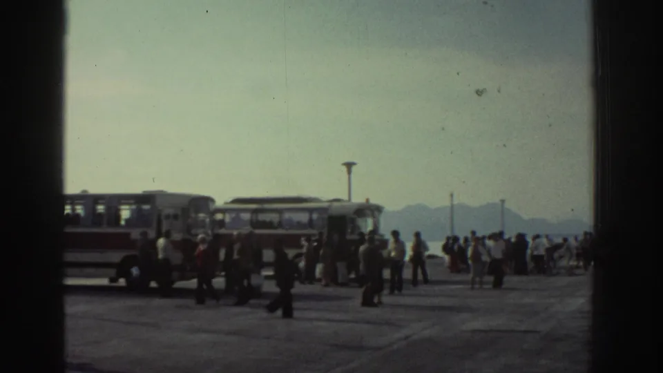 Dockside crowd scene at a Greek ferry port circa 1976, showing passengers, period buses, a tall lamp post, and hazy mountains on the horizon.