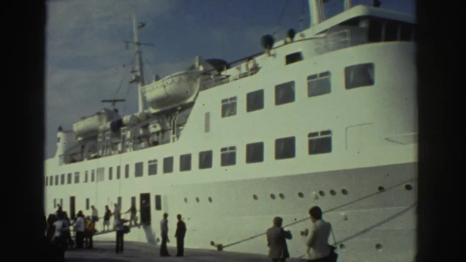 Broadside view of a white multi-deck passenger vessel at a Greek port circa 1976, showing lifeboat davits, multiple cabin-window decks, funnel, and passengers on the pier below.