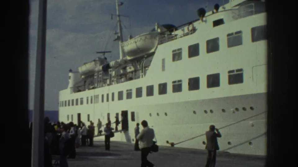 Wide dockside view of a large white passenger ship at a Greek port circa 1976, with a crowd of passengers on the pier; a person photographing is visible in the right foreground.