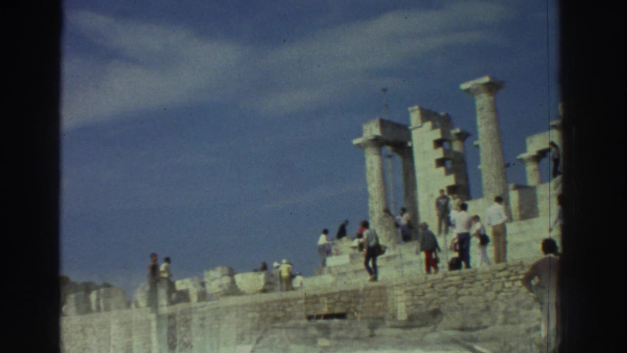 Group of tourists walking among ancient Greek stone ruins and Doric columns on a raised platform under a blue sky with scattered clouds.
