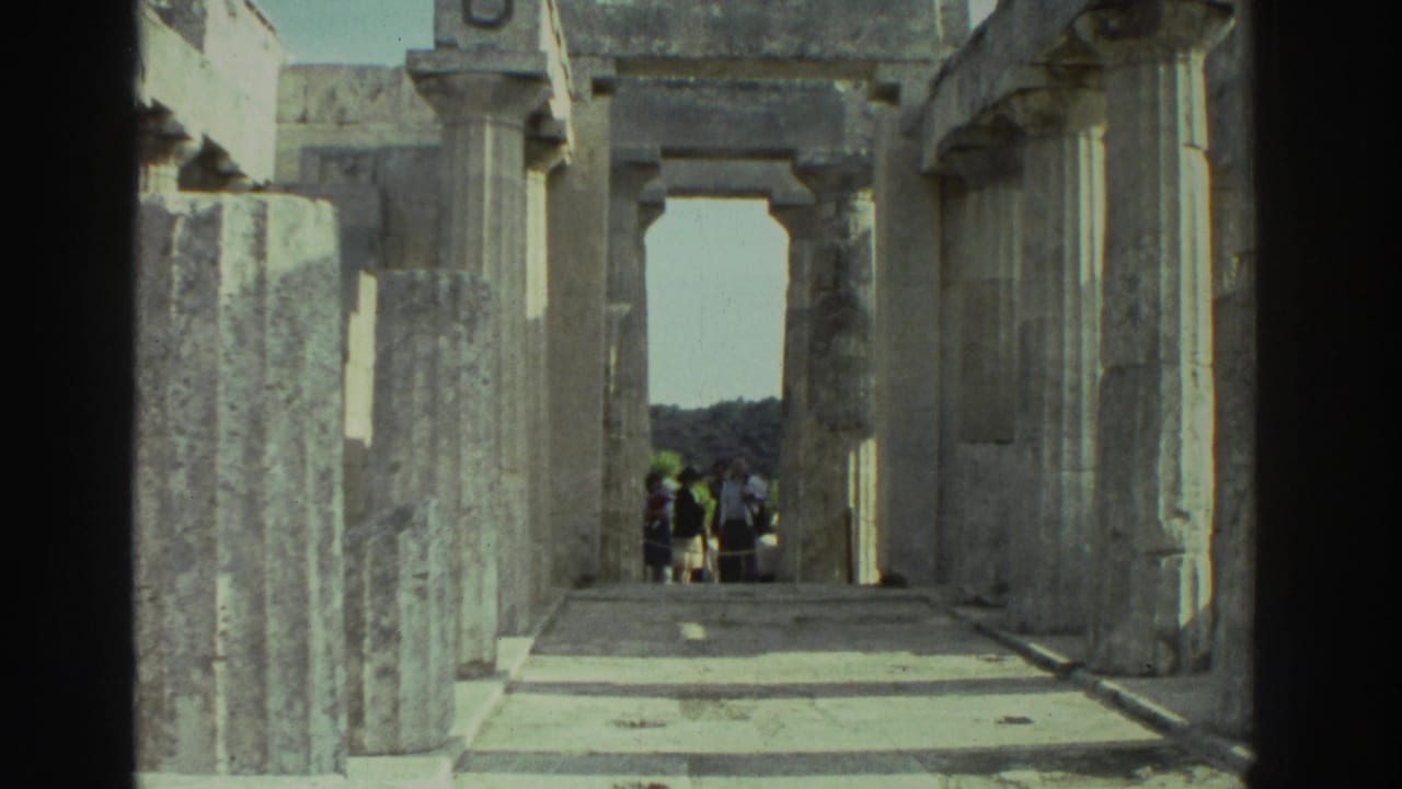 1976 8mm home movie: looking through the interior corridor of an ancient Greek temple, rectangular stone pillars flanking a bright doorway, a small group of tourists visible in the far distance.