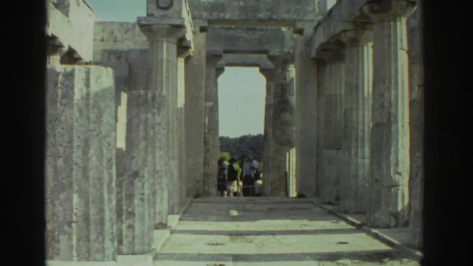 1976 8mm home movie: looking through the interior corridor of an ancient Greek temple, rectangular stone pillars flanking a bright doorway, a small group of tourists visible in the far distance.