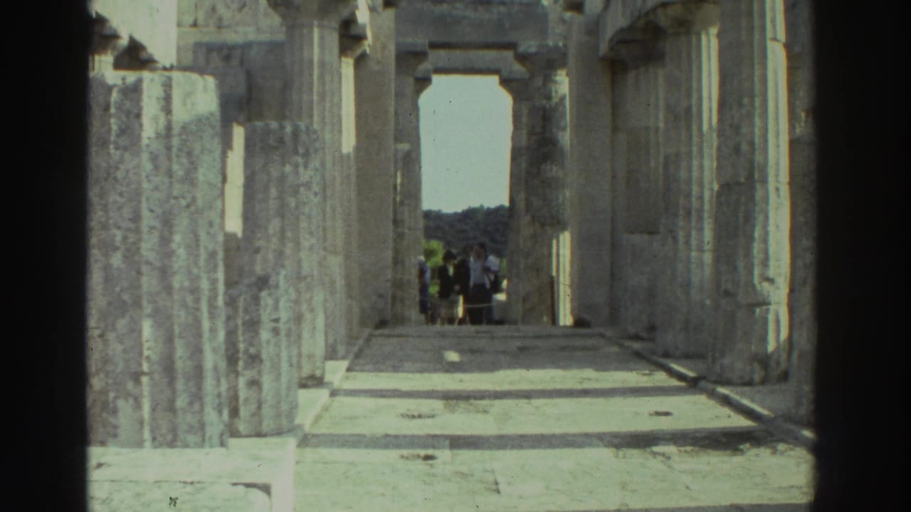 1976 8mm: Greek ancient temple interior viewed through stone columns toward a rectangular doorway; a small group of visitors stands near the opening against a blue sky and wooded hillside.