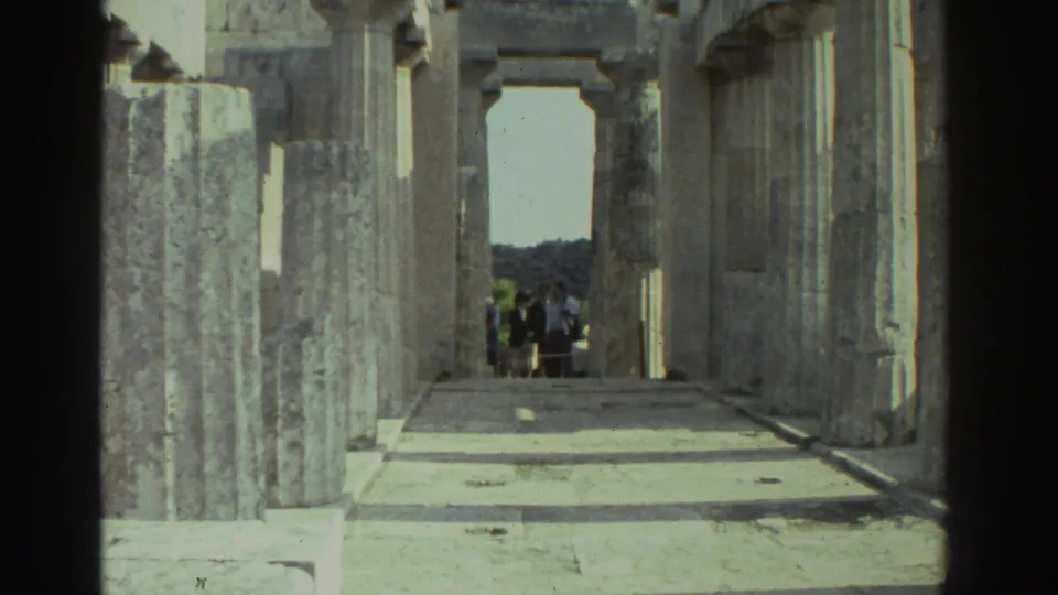 1976 8mm: Greek ancient temple interior viewed through stone columns toward a rectangular doorway; a small group of visitors stands near the opening against a blue sky and wooded hillside.