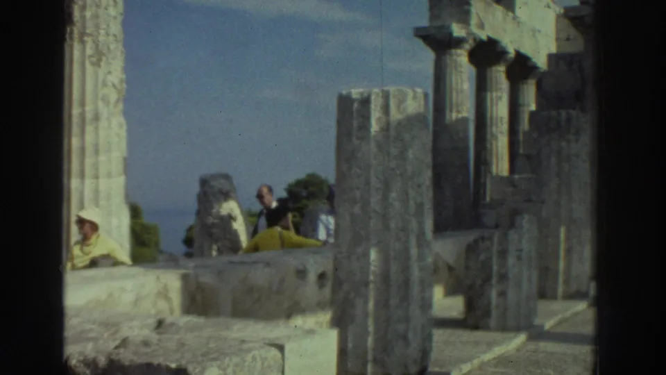 8mm film frame, Greece 1976: Doric columns with tourists in 1970s attire, including a yellow head covering, seated on ancient stone ruins under blue sky