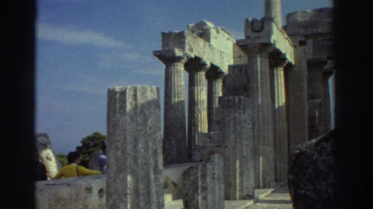 8mm film frame, Greece 1976: low-angle pan across standing Doric columns with stone rubble at base, blue sky with thin clouds, and trees visible beyond