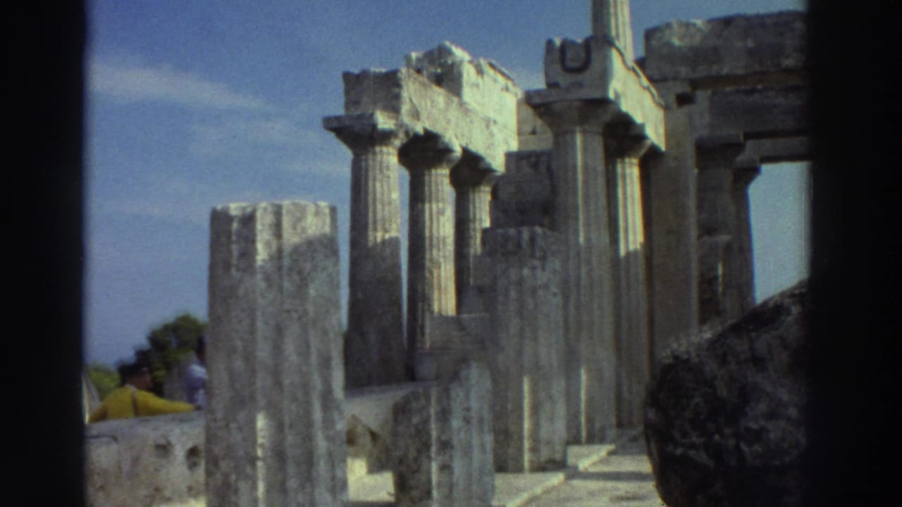 8mm film frame, Greece 1976: cluster of Doric columns under clear blue sky, stone steps and yellow clothing visible at base, trees in background
