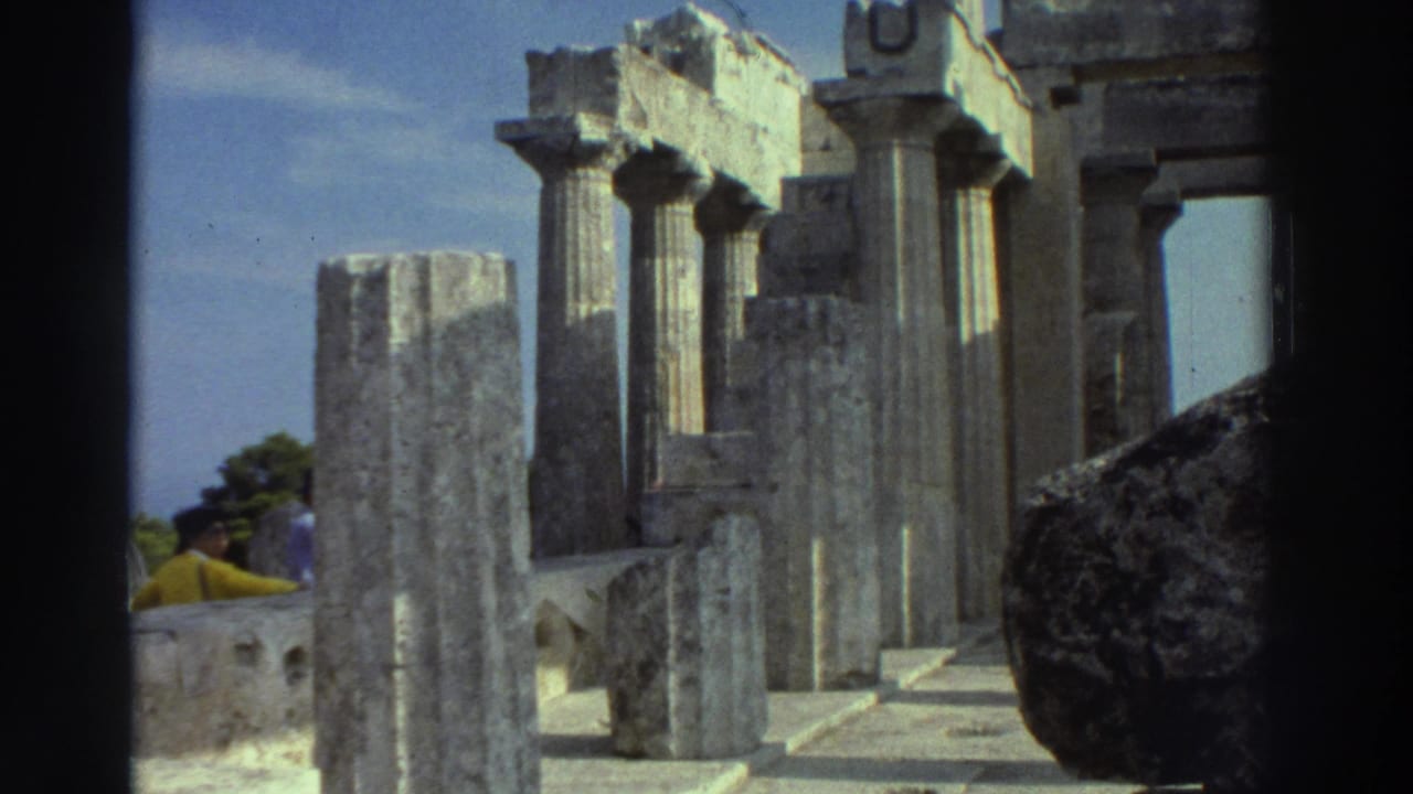 Ancient Greek temple ruins with tall fluted columns under a blue sky; a woman in yellow is visible in the left background.