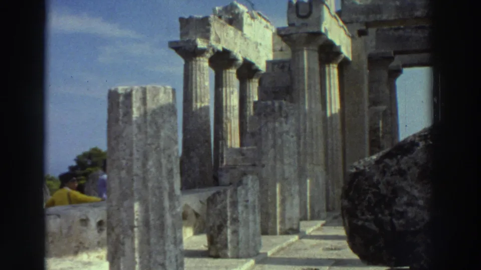 8mm home movie, circa 1976 Greece: fluted stone columns of a classical temple, weathered stone steps and pale limestone blocks, bright blue sky; a tourist in yellow visible in background.