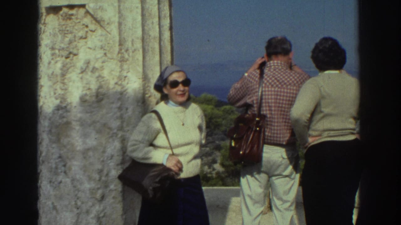 Woman in white sweater, blue headscarf, and dark sunglasses smiling at camera; man in red checkered shirt and a second figure stand behind at a stone overlook.