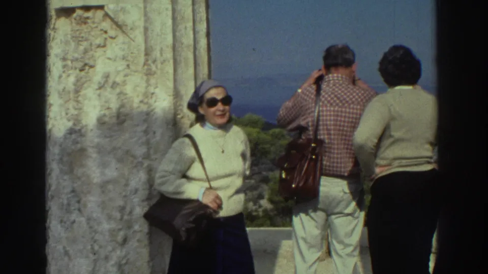8mm home movie, circa 1976 Greece: woman in 1970s dress smiling for camera at ancient stone overlook; man and companion behind her look out toward distant sea and landscape.