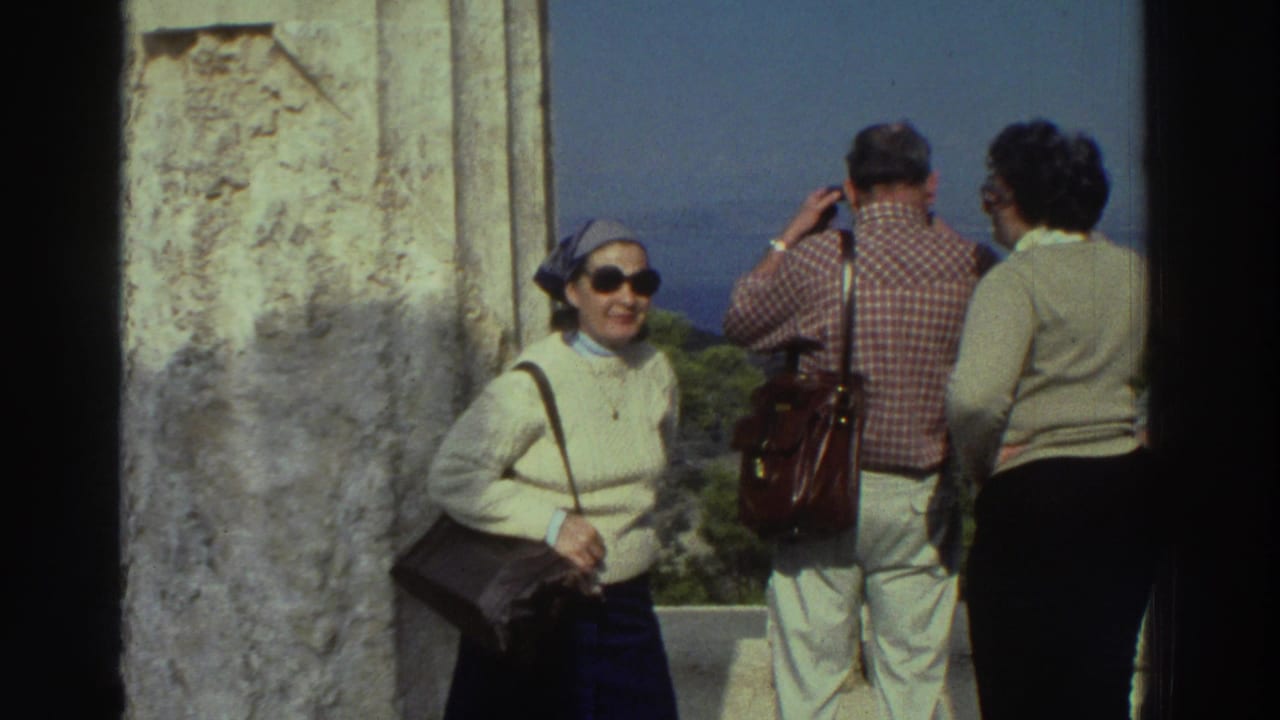 8mm home movie, circa 1976 Greece: tourist woman smiles broadly toward camera as two companions gaze out at the sea from an ancient stone overlook; treeline visible below.