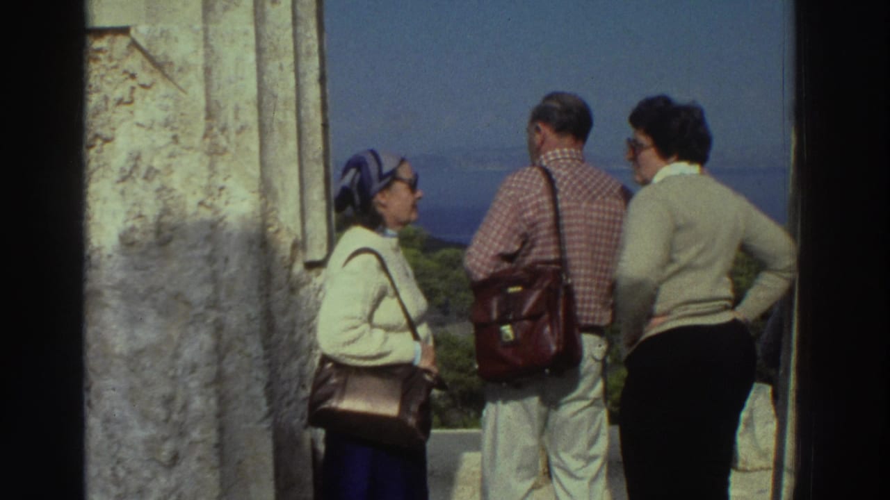 Three tourists in 1970s clothing at an ancient Greek column overlook: woman in blue headscarf, man in plaid shirt with satchel, woman in cream sweater, Greece, 1976.