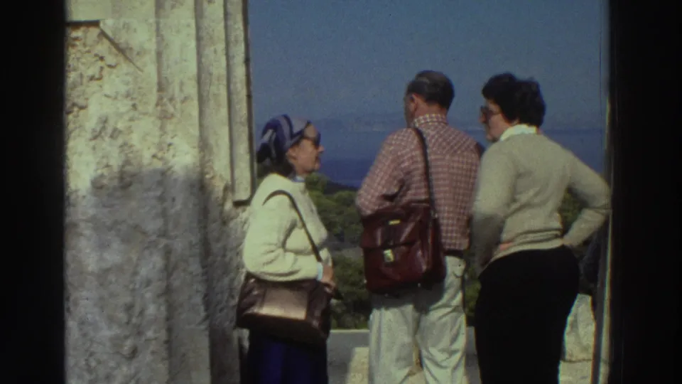 Three tourists in 1970s clothing at an ancient Greek column overlook: woman in blue headscarf, man in plaid shirt with satchel, woman in cream sweater, Greece, 1976.