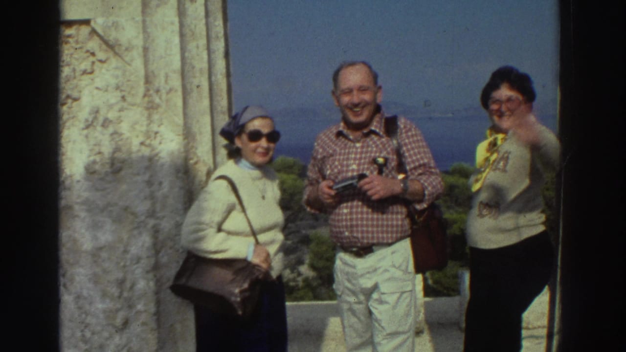 Three tourists facing camera smiling and waving beside an ancient stone column, blue sea in background.