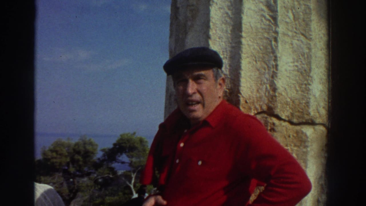 Close view of an older man in a red shirt and dark flat cap smiling near a large ancient stone column, Greece, 1976, with green trees and blue sky visible behind him.