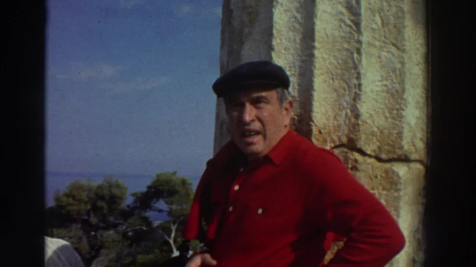 Close view of an older man in a red shirt and dark flat cap smiling near a large ancient stone column, Greece, 1976, with green trees and blue sky visible behind him.