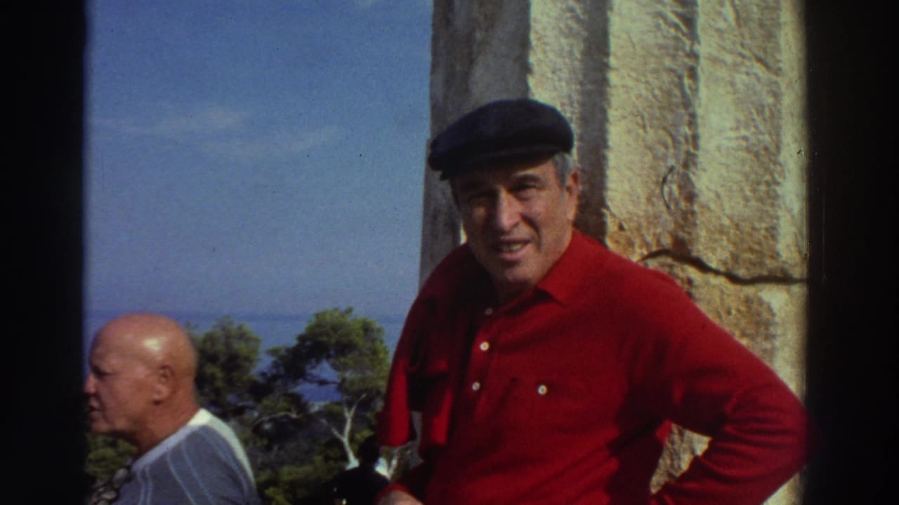 Older man in red shirt and dark flat cap smiling directly at camera beside an ancient stone column, blue sea and green trees visible in background, Greece, 1976.