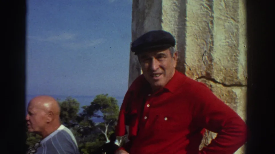 Older man in red shirt and dark flat cap smiling directly at camera beside an ancient stone column, blue sea and green trees visible in background, Greece, 1976.