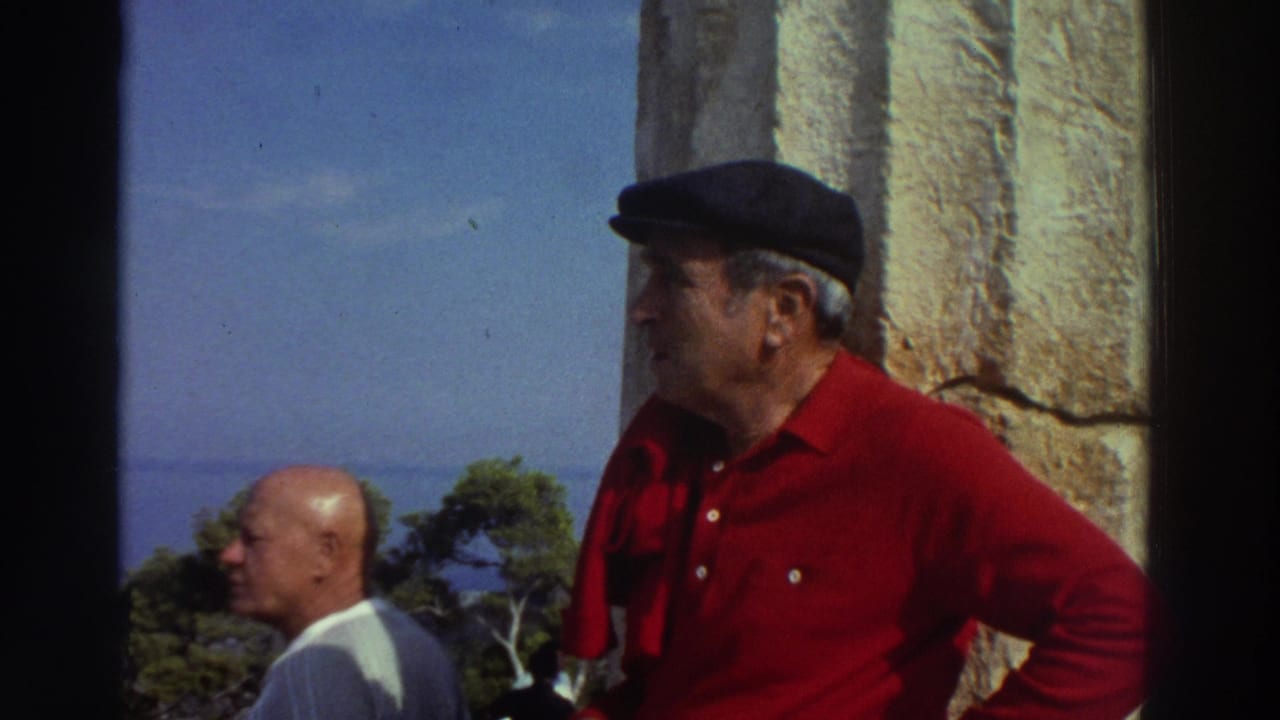 Two men at ancient ruins in Greece, circa 1976 — man in dark beret and red shirt faces left beside a tall stone column; pine trees and sea visible behind.