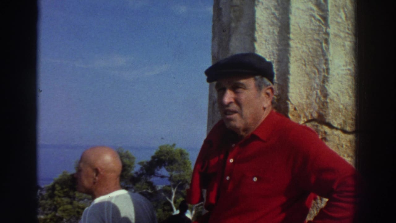 Man in dark flat cap and red shirt turns toward camera appearing to speak; bald companion and weathered stone column visible behind him.