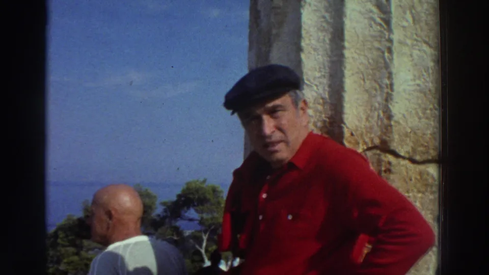 Close framing on a man in a dark beret and red long-sleeve shirt at ancient ruins in Greece, circa 1976; a weathered stone column fills the right side of frame.