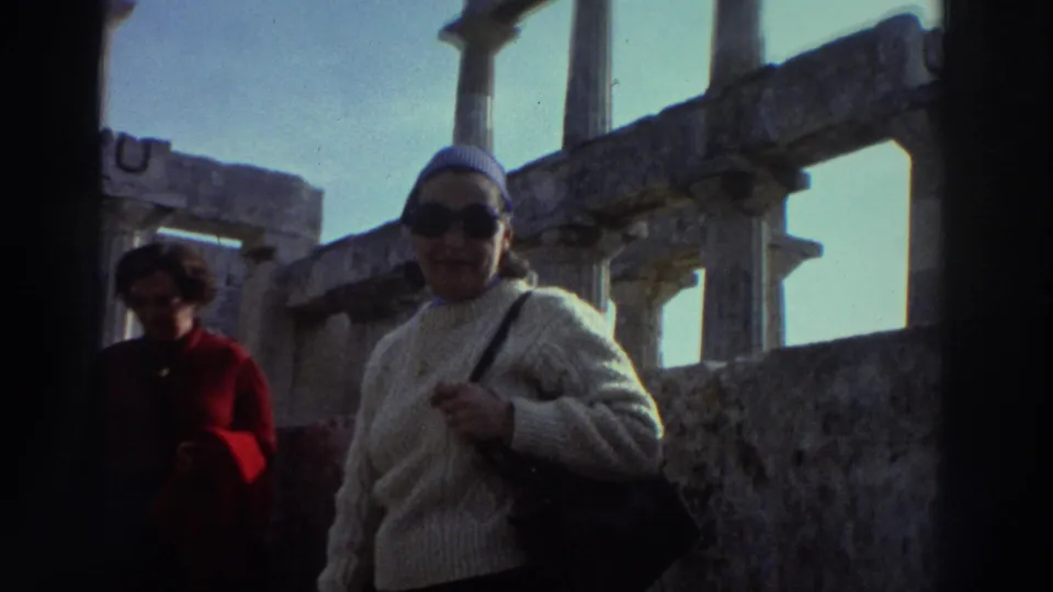 A woman in a cream knit sweater, sunglasses, and light headscarf walks through ancient ruins in Greece, circa 1976; columns and stone walls fill the background with other visitors behind.