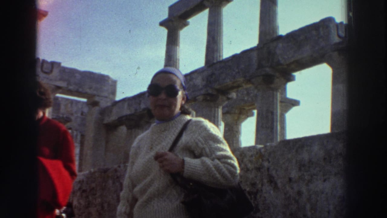 Woman tourist in mid-1970s dress poses beside weathered Greek classical columns at an unidentified ancient site, circa 1976; another visitor in red jacket at frame left.