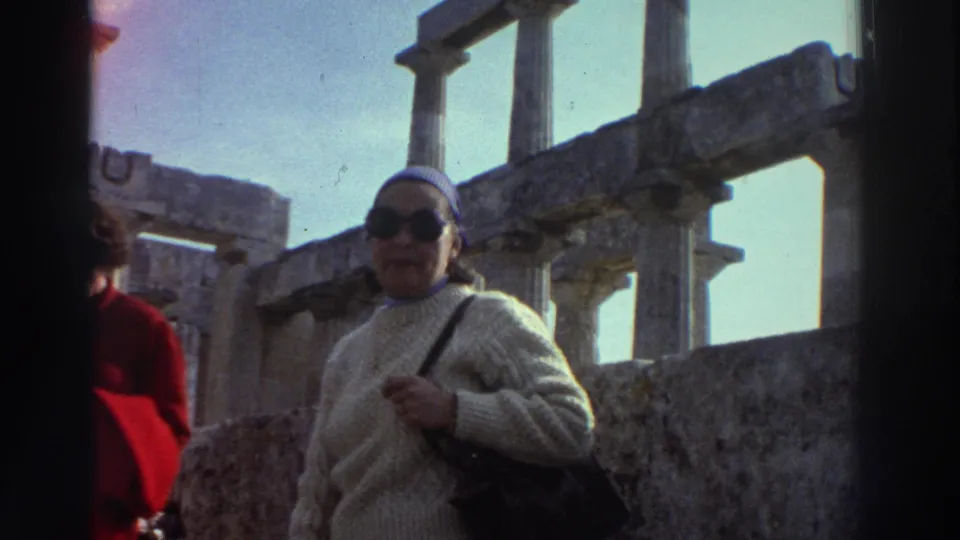 Woman tourist in mid-1970s dress poses beside weathered Greek classical columns at an unidentified ancient site, circa 1976; another visitor in red jacket at frame left.