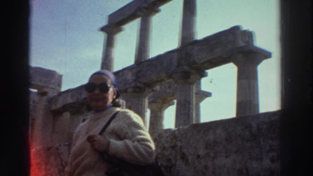 Same woman in cream sweater and blue headscarf faces camera beside ancient stone post-and-lintel columns under clear blue sky.