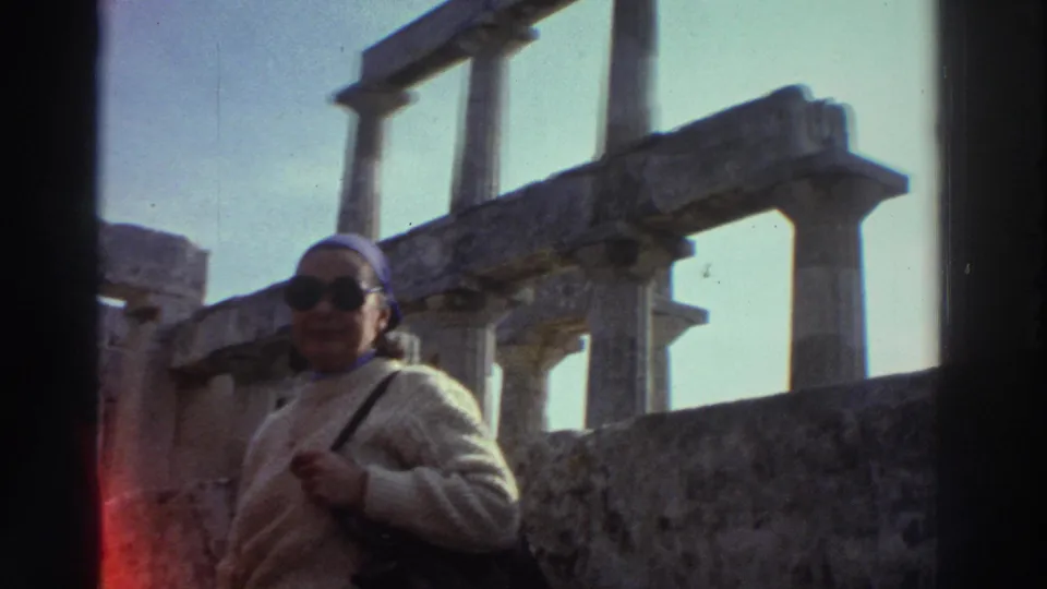 Close shot of woman tourist at Greek ancient ruins, circa 1976, with post-and-lintel columns prominent in background; reddish film artifact visible at right frame edge.