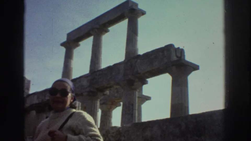 Tourist at unidentified Greek classical ruins, circa 1976, looking upward; weathered stone post-and-lintel columns stand against a clear blue sky.