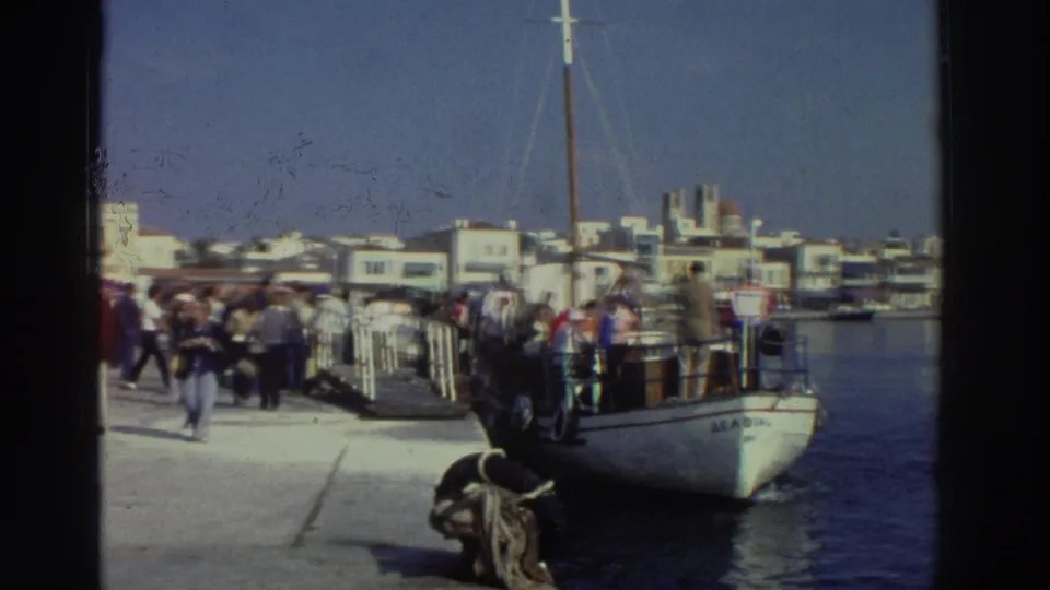 8mm footage of a Greek port waterfront, circa 1976; a moored white boat with partial name lettering at dock, crowds along the quay, whitewashed buildings, and blue sky.