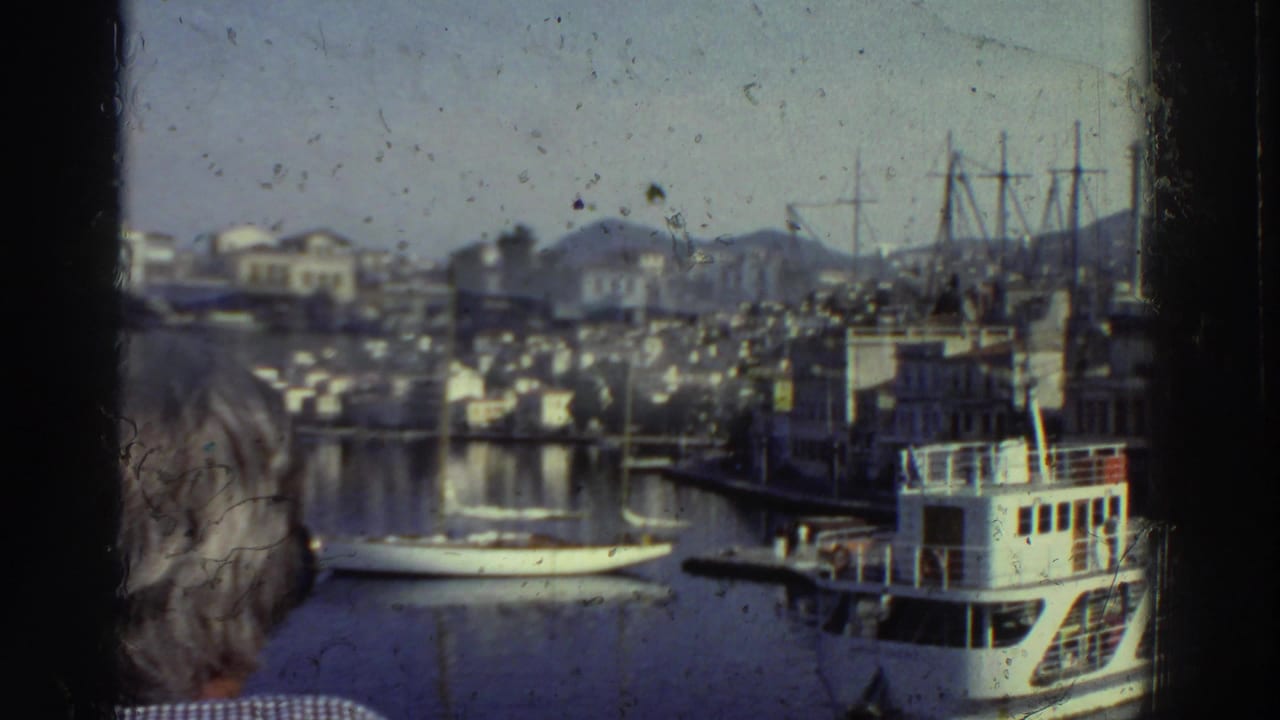 White passenger ferry at anchor in a Greek harbor, 1976; dense white hillside buildings and sailboat masts visible, shot through a window frame.