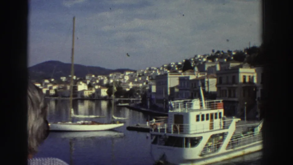 White passenger ferry in Greek harbor, 1976; hillside town with white buildings and sailboat mast behind, partial foreground figure at frame edge.