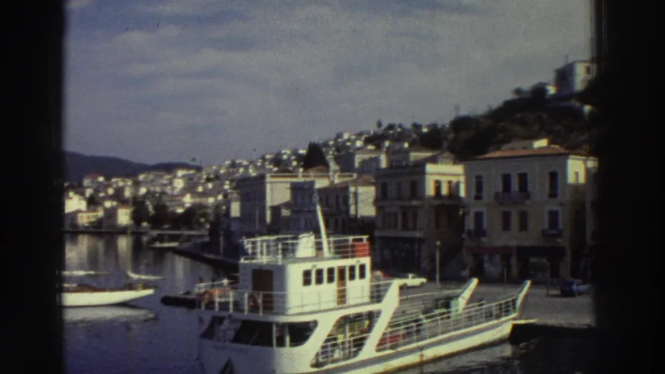 White passenger ferry moored at quay, Greek harbor, 1976; Mediterranean-style multi-story buildings and tree-covered hillside behind.