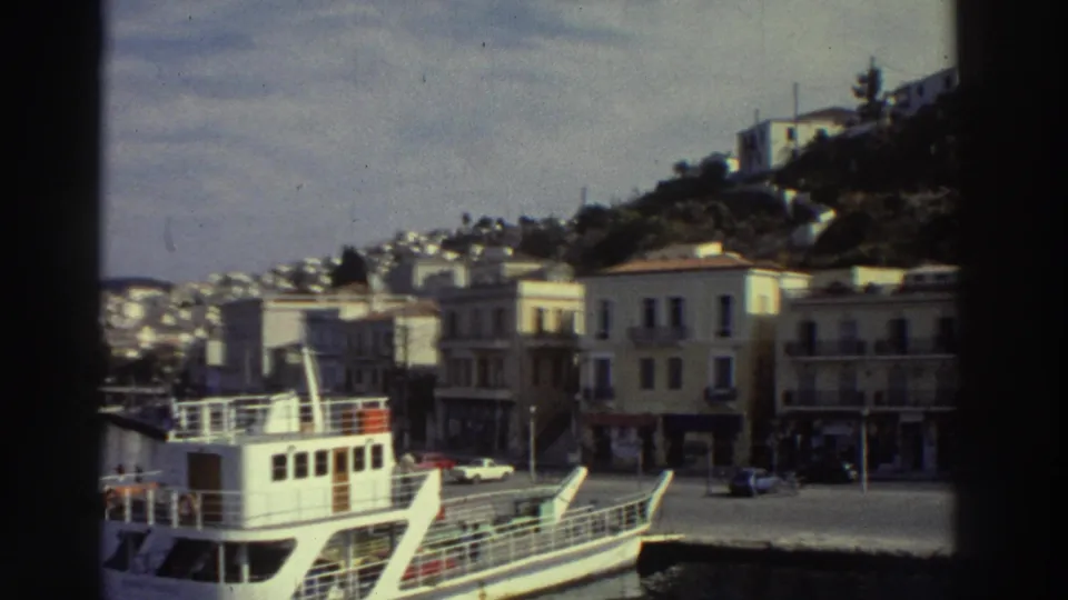 White passenger ferry at dock with yellow-cream Mediterranean waterfront buildings and hillside town, Greek harbor circa 1976.