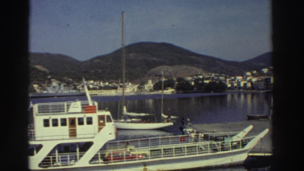 White passenger ferry and smaller white boat moored at a Greek harbor, hillside town and green hills visible across the bay, circa 1976.