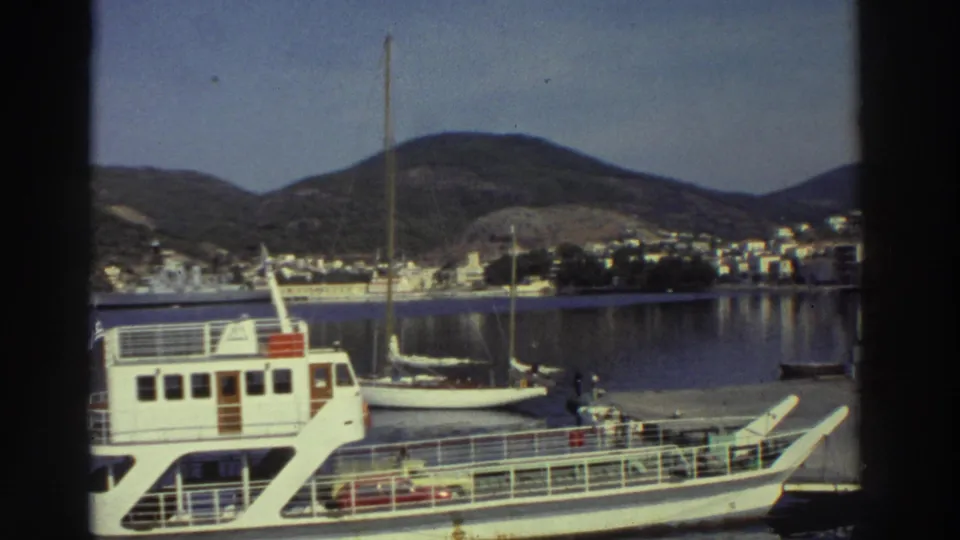 White passenger ferry and smaller white boat moored at a Greek harbor, hillside town and green hills visible across the bay, circa 1976.