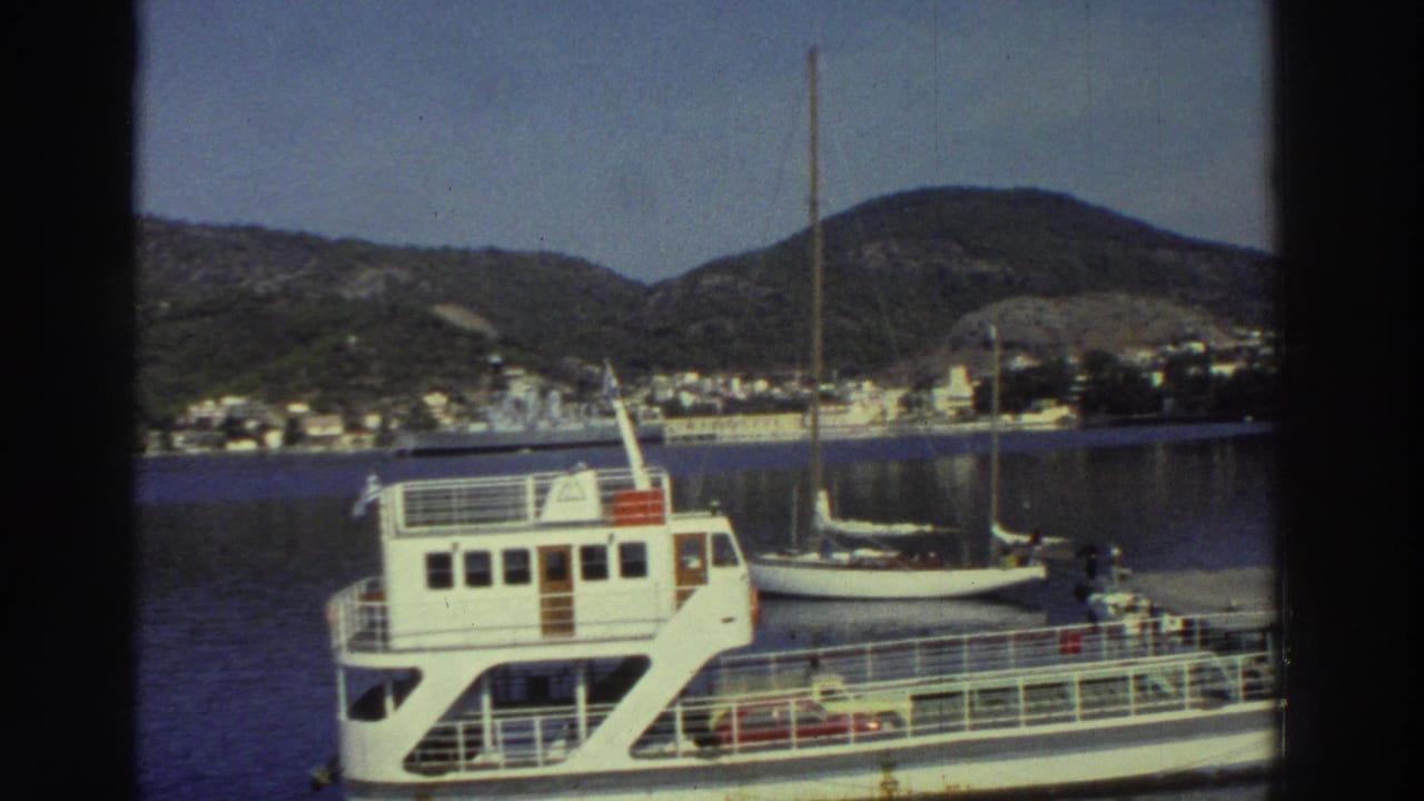 Close view of a white passenger ferry at a Greek harbor, sailboat moored alongside, white-building hillside town across the bay, circa 1976.
