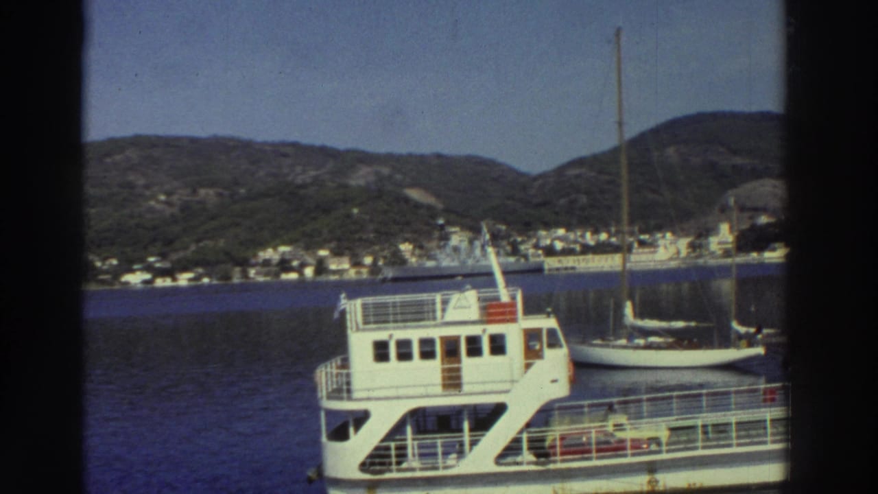 White passenger ferry and companion sailboat at a Greek harbor dock, hillside town of white buildings visible across the bay, circa 1976.