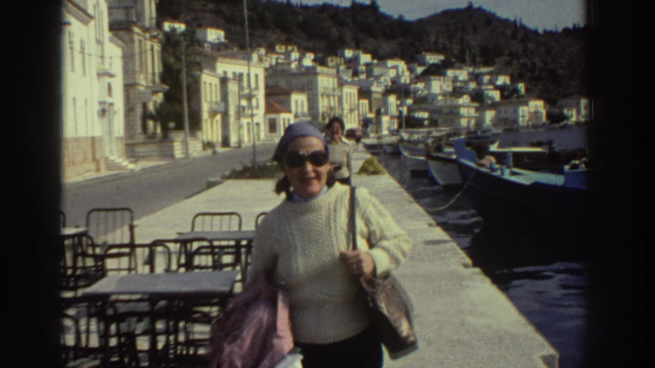 Woman in cream cable-knit sweater and blue headscarf walks along a Greek harbor promenade with cafe tables and moored boats.