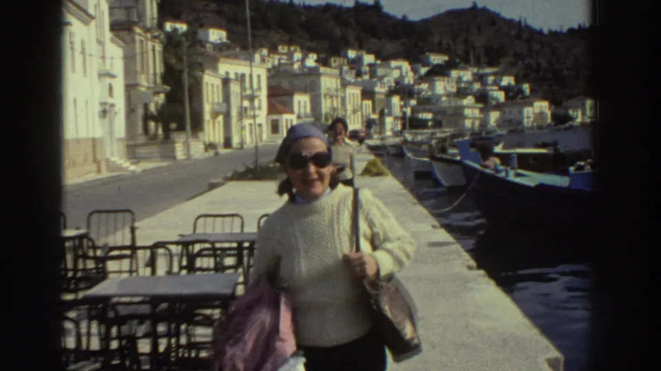 Woman in cream sweater and blue headscarf walks toward camera on a Greek harbor promenade, 1976; blue fishing boats and hillside town behind her.